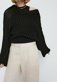 Zwart gebreide trui met een losse pasvorm, voorzien van een wijde halslijn en een textuurpatroon. Gecombineerd met lichtbeige, op maat gemaakte broeken.