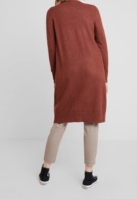 Pitkä, ruosteenruskea neuletakki, valmistettu kevyestä materiaalista ja väljällä istuvuudella. Päällä kuvioidut, lyhyet housut ja mustat lenkkarit.