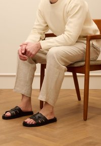 Black leather sandals with multiple straps, flat sole, worn with light beige linen trousers and a cream sweater, seated on a wooden chair.