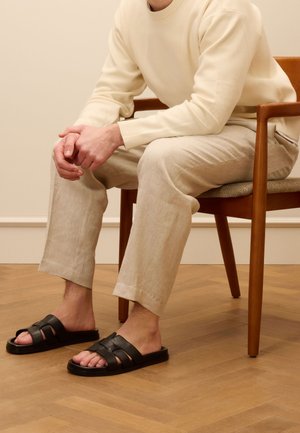 Schwarze Leder-Sandalen mit mehreren Riemen, flache Sohle, getragen mit hellbeigen Leinenhosen und einem cremefarbenen Pullover, sitzend auf einem Holzstuhl.