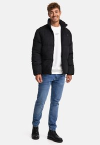 Sort puffjakke med høj krave, tekstureret stof, lynlås lukning og kontrasterende hvid sweatshirt; parret med lyseblå jeans og sorte støvler.