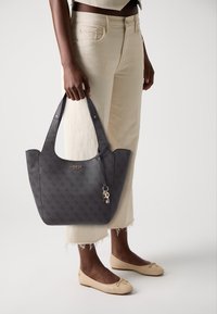 Borsa a spalla grigia a motivo con manici doppi e un charm, tenuta da una persona con pantaloni chiari e ballerine beige. Sfondo semplice.