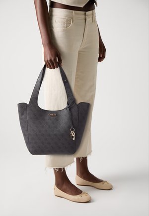 Borsa a spalla grigia a motivo con manici doppi e un charm, tenuta da una persona con pantaloni chiari e ballerine beige. Sfondo semplice.