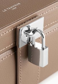 Caja de almacenamiento de cuero marrón con un candado y cierre de metal plateado, con costuras visibles y una textura suave. Marca grabada en la tapa.