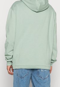 Felpa verde menta con cappuccio, maniche lunghe e polsini a costine, abbinata a jeans chiari strappati con tasca a toppa.