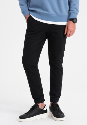 Zwarte joggingsbroek van zachte stof, met een elastische tailleband en boorden. Draagt het met zwarte sneakers met witte accenten.