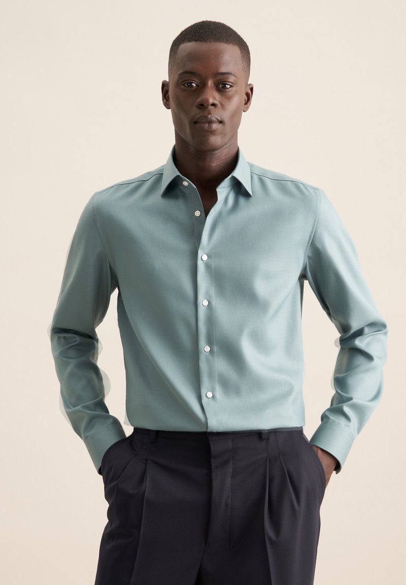 Jasnoniebieska koszula zapinana na guziki o gładkiej fakturze, z długimi rękawami i białymi guzikami, w zestawieniu z ciemnymi spodniami. Prosty, klasyczny design.