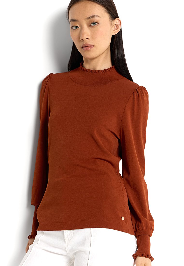 Einer rostfarbenen Langarmbluse aus einem weichen Stoff, mit einem hohen Kragen und Rüschendetails sowie gepufften Schultern, kombiniert mit weißen Hosen.