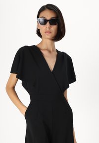 Svart jumpsuit med dyp V-hals og lette ermer. Glatt tekstur, tilpasset passform og sidelommer. Kombinert med stilige svarte solbriller.