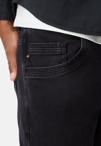 Sorte denim jeans med en glat tekstur, der har en sidelomme og guld hardware accenter. Synlige stikningsdetaljer langs kanterne.