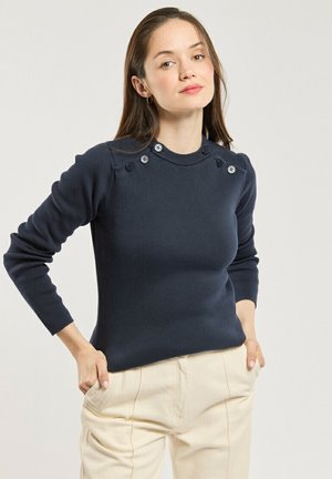 Femme portant un pull bleu marine foncé boutonné et un pantalon beige clair, debout les mains dans les poches sur un fond clair uni.