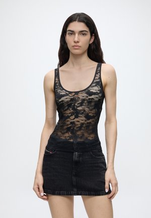 Donna con lunghi capelli scuri indossa un top in pizzo nero trasparente e una gonna corta di jeans nero, in piedi contro uno sfondo chiaro e uniforme.