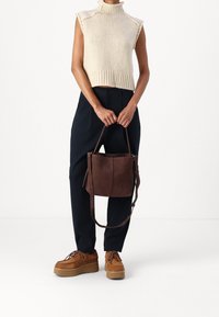 Bolso de tote de ante marrón con asa para el hombro, diseño sencillo, textura suave, combinado con una parte superior sin mangas de punto crema y pantalones oscuros.