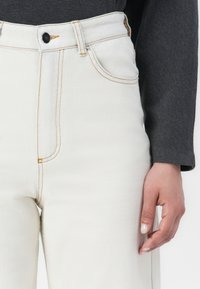 Høyt liv, hvite denimjeans med mørk søm. Har frontlommer, knappelukking foran og en tettsittende silhuett.