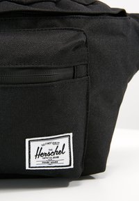 Černá hip taška Herschel vyrobená z odolné látky, s přední kapsou na zip a bílou značkovou etiketou s černým textem a obrysem.