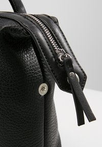 Närbild av en svart texturerad läderväska med silverfärgad dragkedja och metallknapp, försedd med en läderdragflik fäst vid dragkedjan.