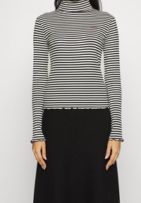Raitapaitainen mustavalkoinen pitkähihainen turtleneck-neule, jossa on aaltoileva helma ja hihansuut, teksturoitu kangas, jossa on pieni brodeerattu logo.