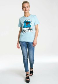 Light blue T-shirt featuring Cookie Monster graphic, black lettering, and a relaxed fit, paired with dark blue skinny jeans and black platform shoes.