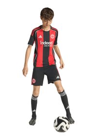 Nogometna oprema s crno-crvenim prugastim dresom, crnim hlačama i čarapama, s Adidasovim brendom i istaknutim logom na majici.
