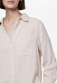 Camisa abotonada de color beige claro, hecha de una tela suave y texturizada. Presenta un cuello clásico y un bolsillo frontal en el lado izquierdo.