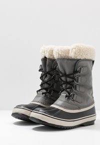 Bottes d'hiver grises avec doublure en fausse fourrure, tissu texturé, semelles en caoutchouc et liseré de couleur claire en contraste. Laçage à l'avant et accents renforcés.