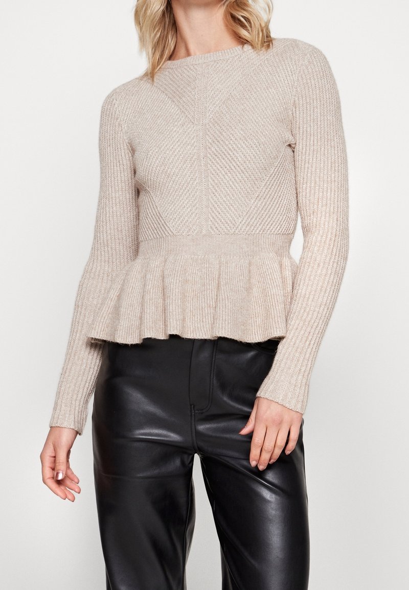 Pull beige en maille avec une texture côtelée, présentant un corsage cintré et un ourlet péplum évasé, associé à un pantalon en cuir noir taille haute.