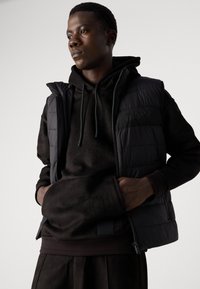 Black padded vest with a quilted design and a zippered front, layered over a black hoodie with a textured finish and kangaroo pocket.
