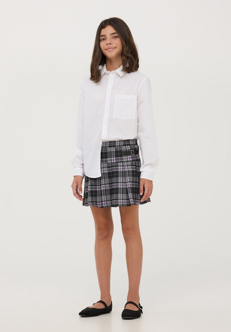 Camicia bianca a bottoni con taschino sul petto, abbinata a una gonna plissettata a quadri nera con accenti viola e bianchi. Ballerine nere.