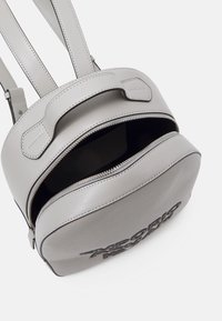 Vaalea harmaa nahkarepulle, jossa on musta vetoketju osittain auki, tekstuuri pinta ja musta kirjailtu logo etutaskussa.