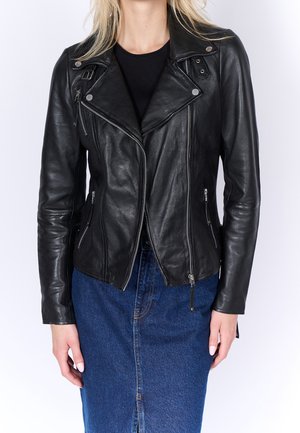 Femme portant une veste de motard en cuir noir avec des poches zippées et une jupe en jean bleue sur un fond blanc uni.