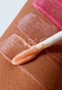 Échantillons de gloss brillant en teintes rose, or rose et transparent appliqués sur la peau avec un pinceau applicateur.
