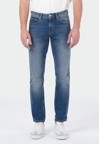 Homme portant un jean bleu délavé et des baskets blanches, se tenant contre un fond uni avec les mains détendues sur les côtés.