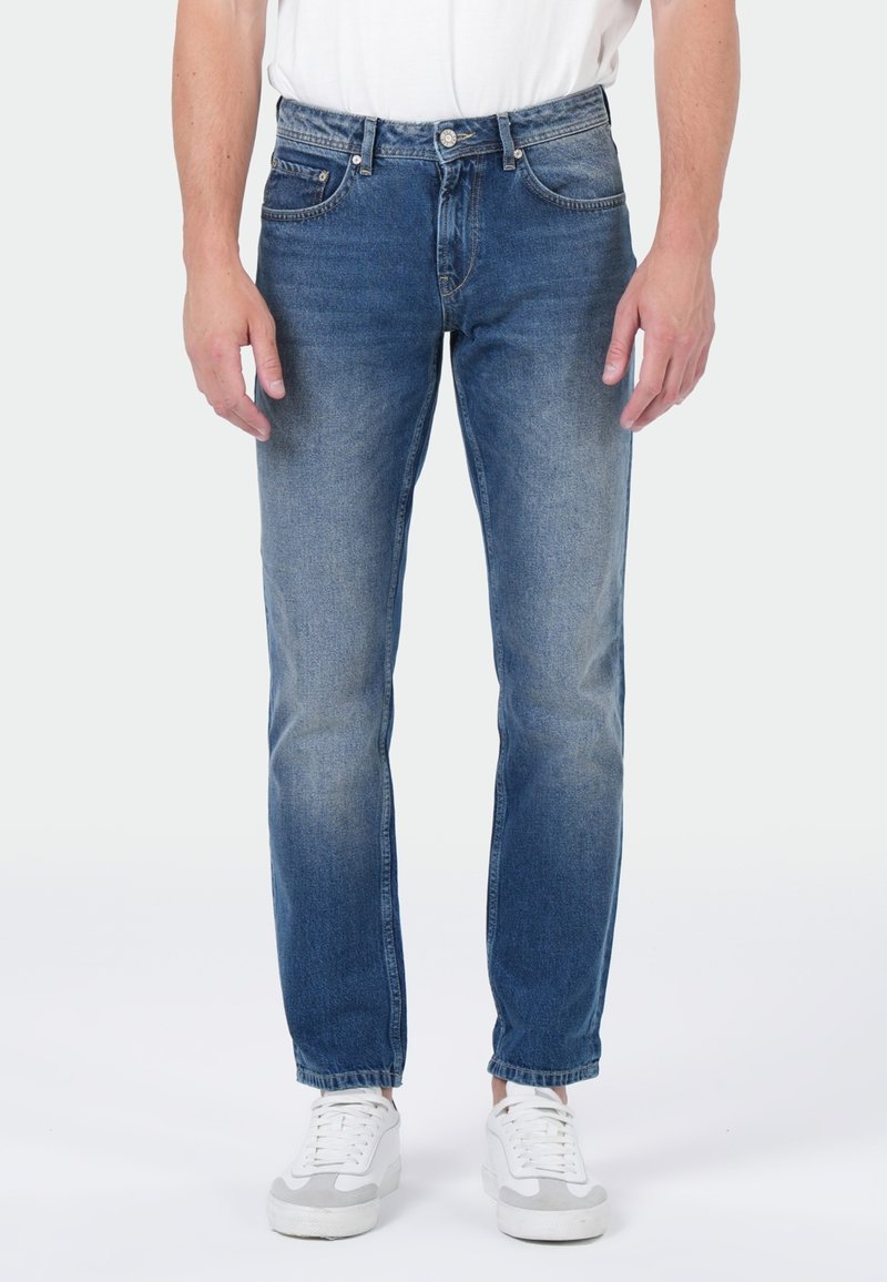 Homme portant un jean bleu délavé et des baskets blanches, se tenant contre un fond uni avec les mains détendues sur les côtés.