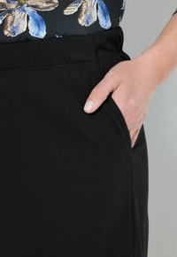 Pantalons noirs à taille élastique avec une poche latérale, en tissu texturé, présentant une finition lisse. Vue rapprochée de la main dans la poche.