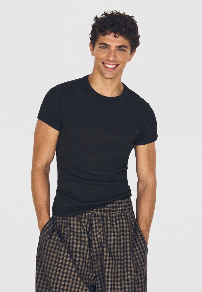 Young man smiling, wearing a fitted black short-sleeve shirt and loose, checkered brown and black pants with hands in pockets.