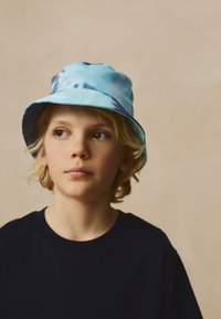 Svetlomodrá vedierková čiapka s tie-dye vzorom a mäkkou textúrou, vybavená širokým okrajom a okrúhlou korunou, nosená dieťaťom v tmavomodrom tričku.