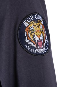 Marineblåt stof med et broderet patch, der viser en brølende tiger og teksten "TOP GUN EN AMERIKANSK IKON" på ærmet.
