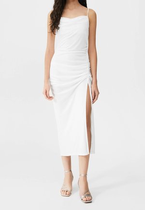 Robe blanche sans manches avec un tissu drapé et un ruché sur le côté. Présente une fente montée à la cuisse et une texture douce, assortie à des sandales argentées à brides.