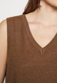 Pull sans manches marron avec décolleté en V, confectionné dans un tissu doux et texturé, présentant un léger éclat et une silhouette ajustée.