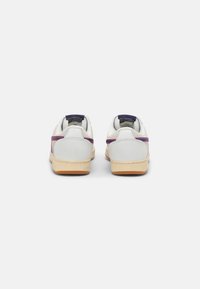 Baskets blanches avec une finition en cuir lisse, des bandes contrastantes rouges et bleues, et une semelle en caoutchouc gomme. Vue arrière montrant le col rembourré et l'étiquette.