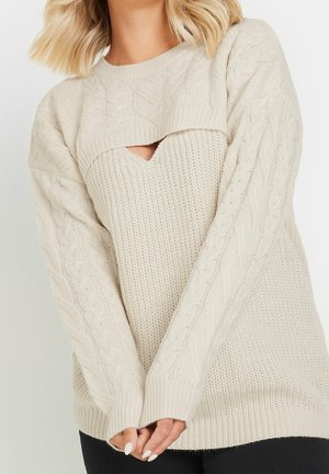 Kvinde iført cremefarvet kabelstrikket sweater med lagdelt design og et lille trekantet udskæring ved brystet, hænderne foldet foran.
