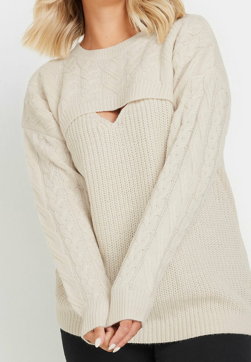Femme portant un pull crème en maille torsadée avec un design à couches et une petite découpe triangulaire au niveau de la poitrine, les mains jointes devant.