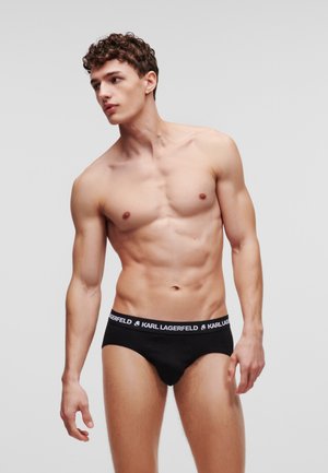 Giovane uomo con capelli ricci che indossa slip neri Karl Lagerfeld, in piedi contro uno sfondo grigio chiaro uniforme, che guarda di lato.