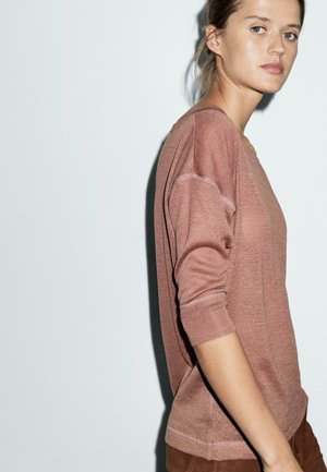 Kvinde med lys hud og blondt hår iført en støvet lyserød striktrøje og brune bukser, stående foran en ensfarvet lys baggrund.