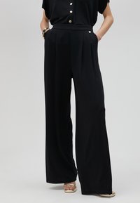 Pantalones de pierna ancha negros, confeccionados en una tela suave, con pliegues frontales, bolsillos laterales y un detalle de cinturón a juego en la cintura.