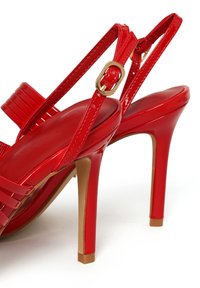Sandalias de tacón alto de charol rojo con tiras al tobillo y hebilla en tono dorado, con tacón de aguja estrecho y diseño de punta abierta.