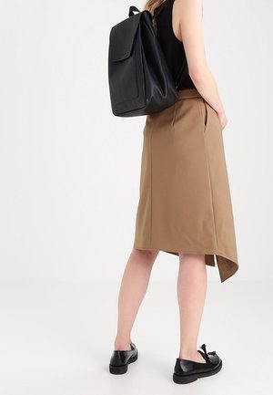 Frau trägt ein schwarzes ärmelloses Oberteil, einen asymmetrischen Rock in Kamelhaarfarbe, schwarze Loafers mit Quasten und einen schwarzen Leder-Rucksack, steht vor einem weißen Hintergrund.