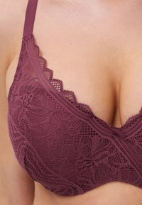 Reggiseno in pizzo malva con bordi a scallop e motivi floreali. Texture morbida e spalline regolabili. Presenta una scollatura a V.