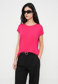 Ροζ μωβ κοντομάνικη μπλούζα από απαλό ύφασμα, συνδυασμένη με μαύρο παντελόνι ψηλόμεσο, με στρογγυλό πόμολο στη ζώνη. Φοράει μαύρα γυαλιά ηλίου.