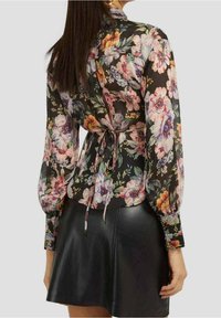 Bluse mit Blumenmuster aus transparentem schwarzem Stoff, mit langen Ärmeln, einer Bindung in der Taille und einem lockeren Schnitt. Kombiniert mit einem schwarzen Lederrock.